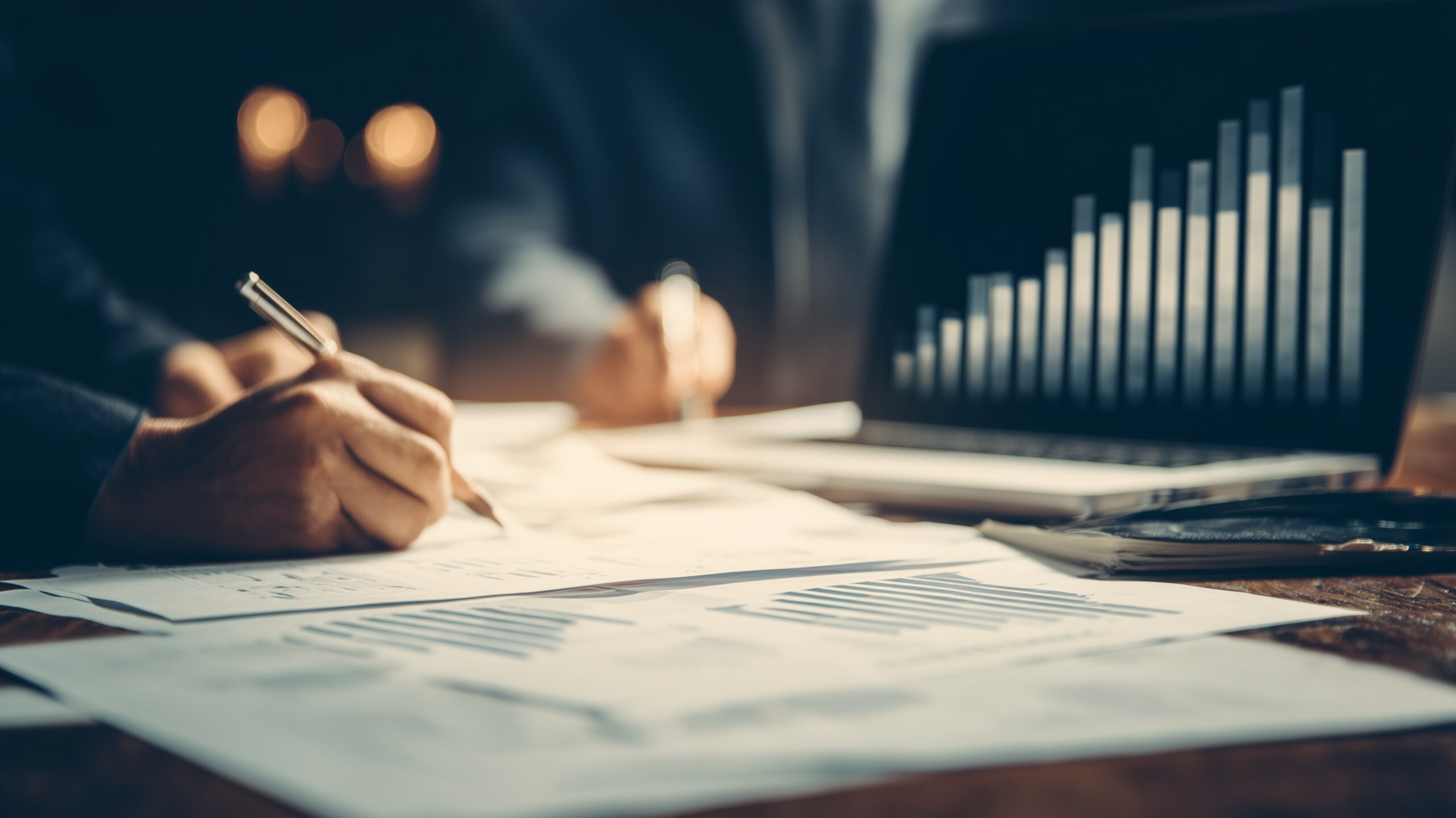 Eine Person notiert etwas auf einem weißen Blatt. Notebook mit Diagramm im Hintergrund