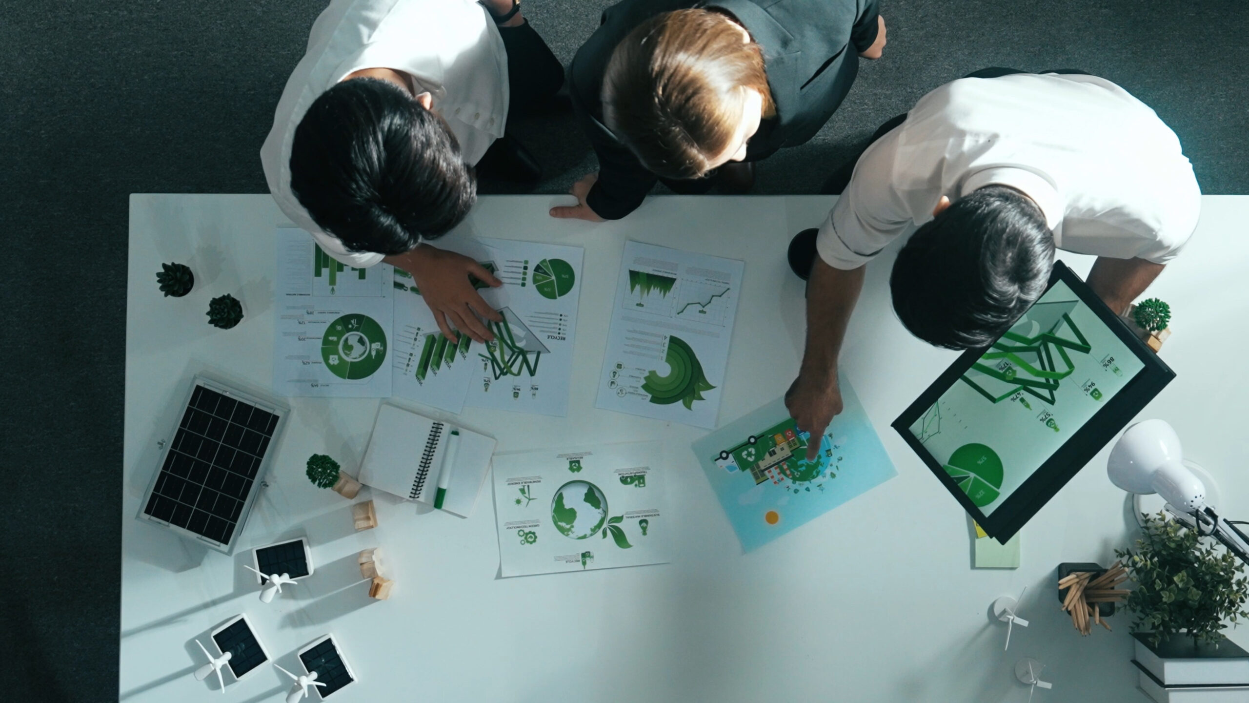 Drei Männer schauen auf Dokumente auf einem Tisch, zwei zeigen auf Schaubilder